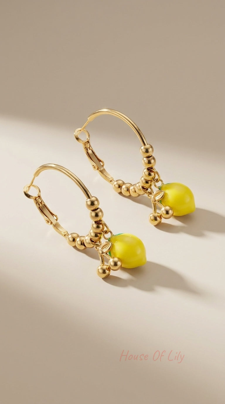 Gold hoop earrings featuring beaded detailing and dangling, glossy yellow lemon charms on a neutral beige background.