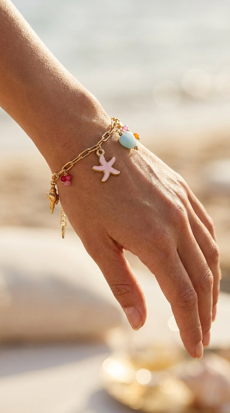 Gold chain bracelet with pink starfish, blue bead, and seashell charms worn on a wrist against a sunny beach background.