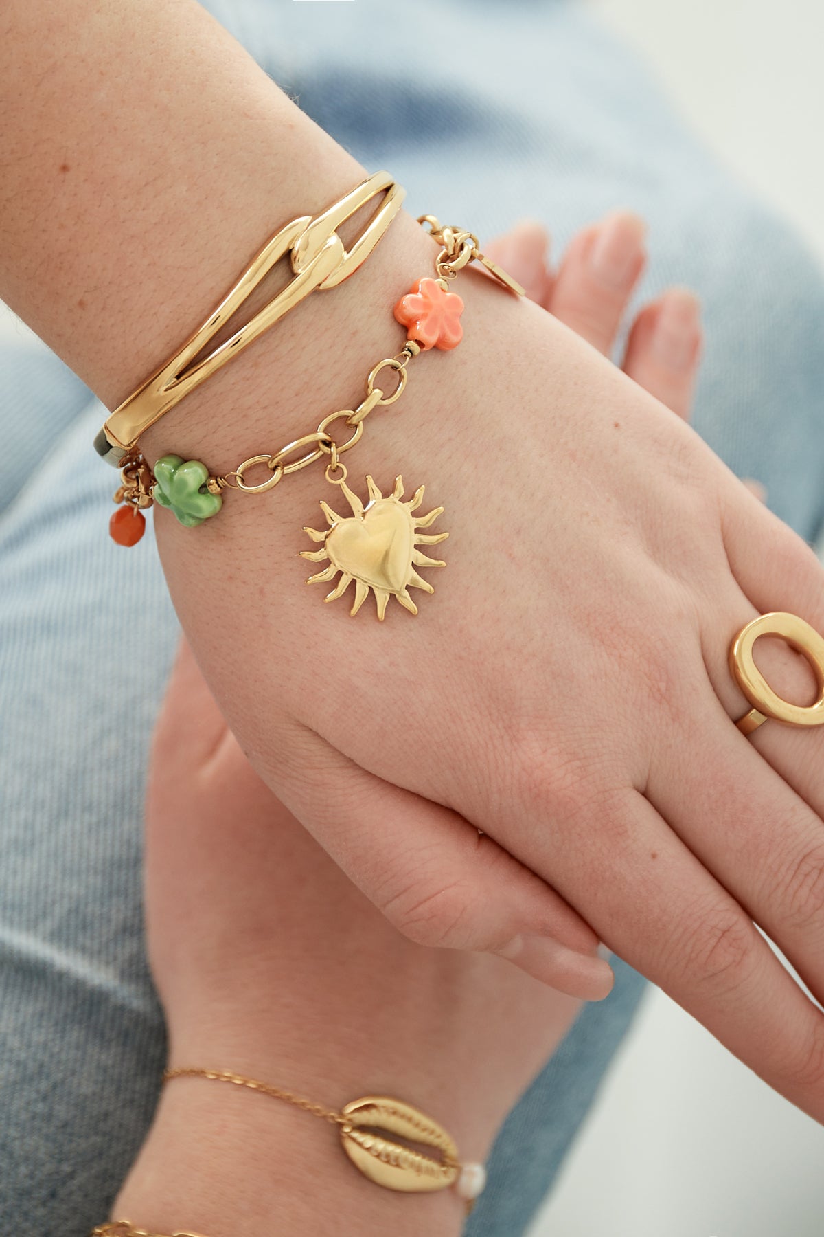 Bracelet worn on wrist styled with gold bangle and ring, showing delicate chain with flower charms and sunburst heart pendant