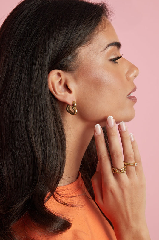 Woman wearing gold heart-shaped hoop earrings and gold rings with styled hair and orange top