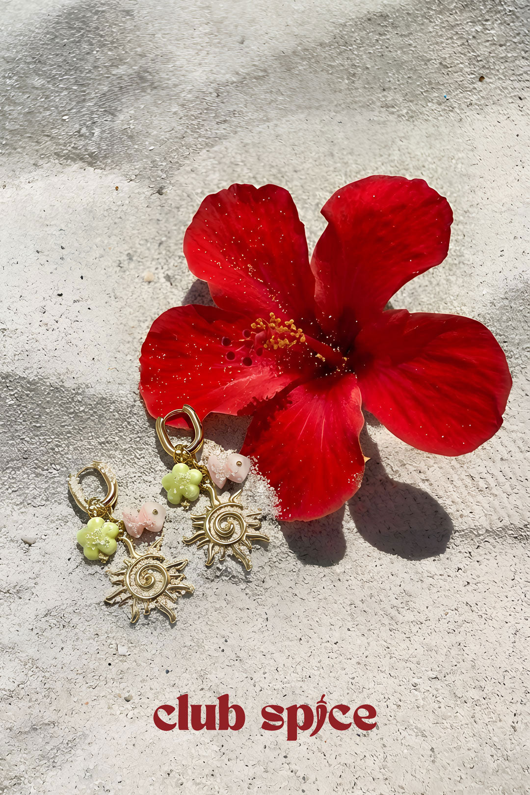 Gold-tone sun charm drop earrings displayed next to a red hibiscus flower on a textured stone surface with Club Spice branding