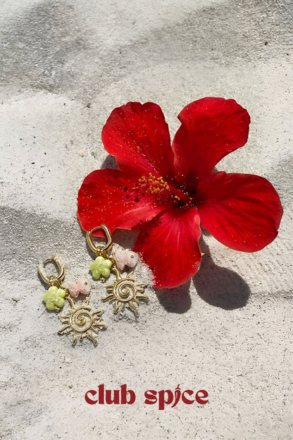 Gold-tone sun charm drop earrings displayed next to a red hibiscus flower on a textured stone surface with Club Spice branding