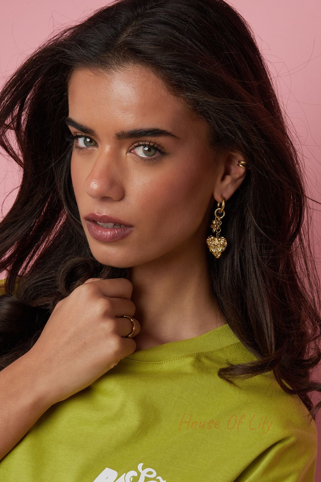 Model wearing gold textured heart drop earring with flower and pearl charms, styled with a green shirt and pink backdrop