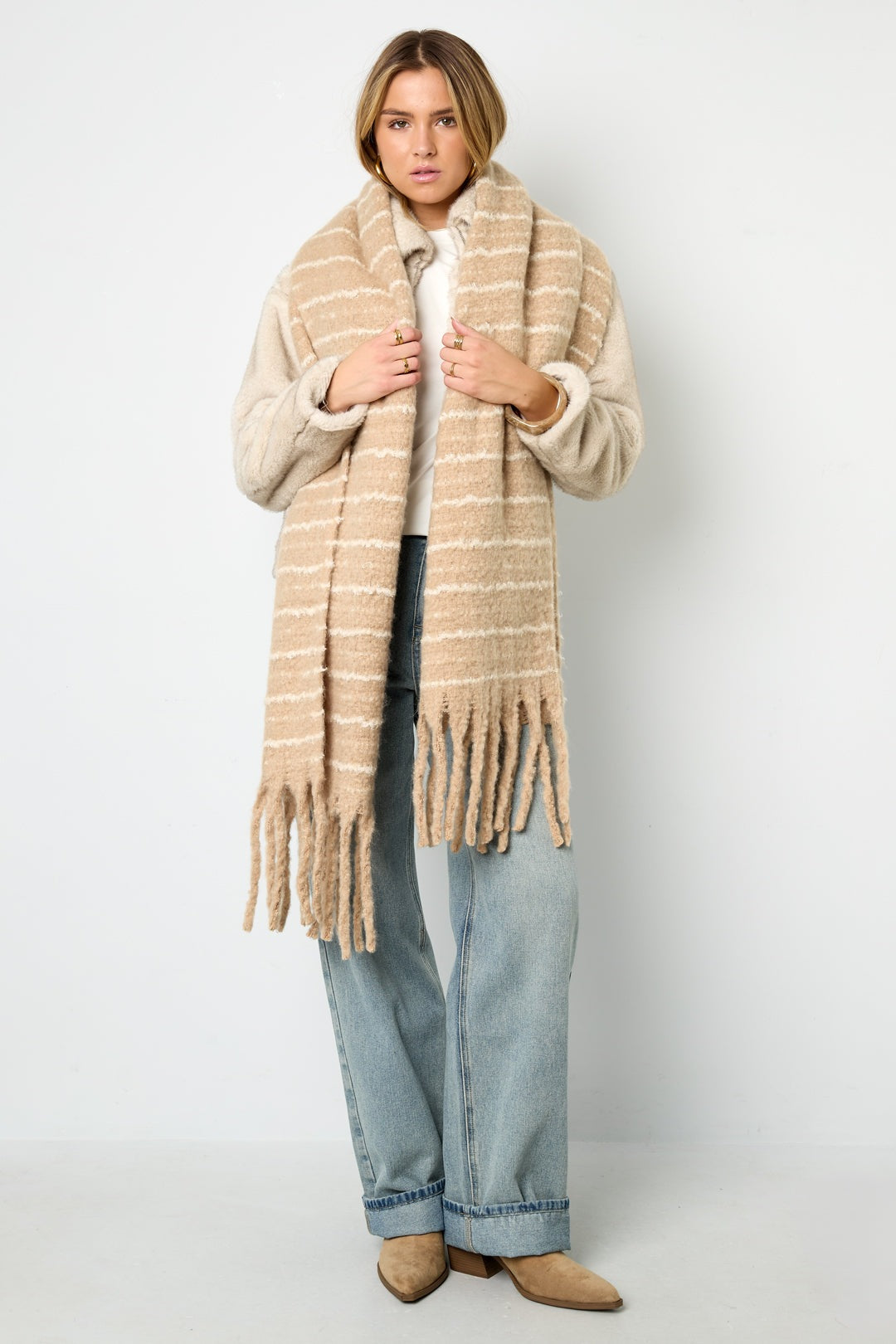 Model wearing beige striped scarf styled over light sweater and blue jeans with tan boots