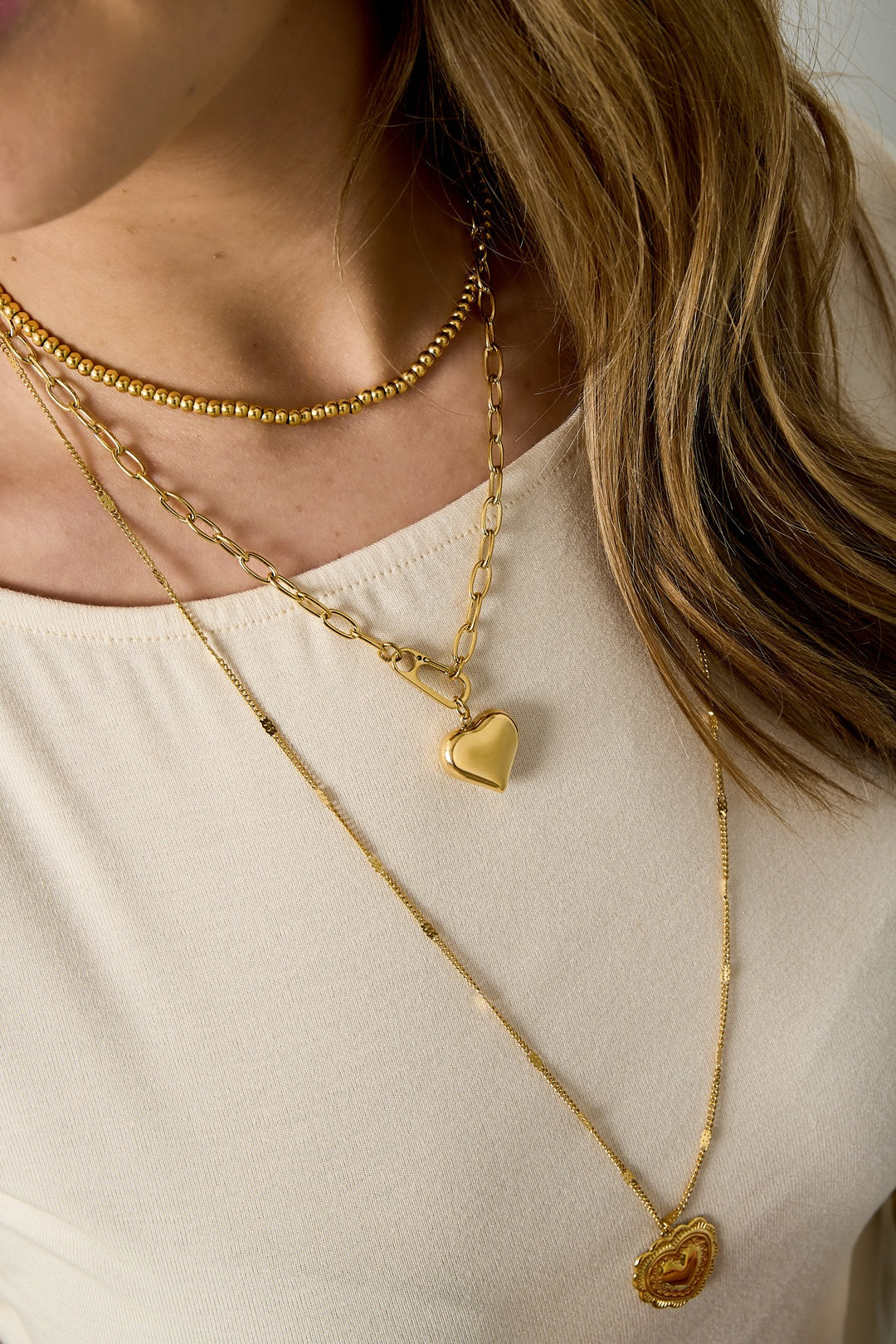 Close-up of woman wearing layered gold necklaces including chunky heart pendant necklace with soft beige top