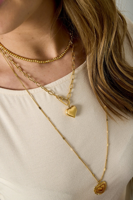 Close-up of woman wearing layered gold necklaces including chunky heart pendant necklace with soft beige top
