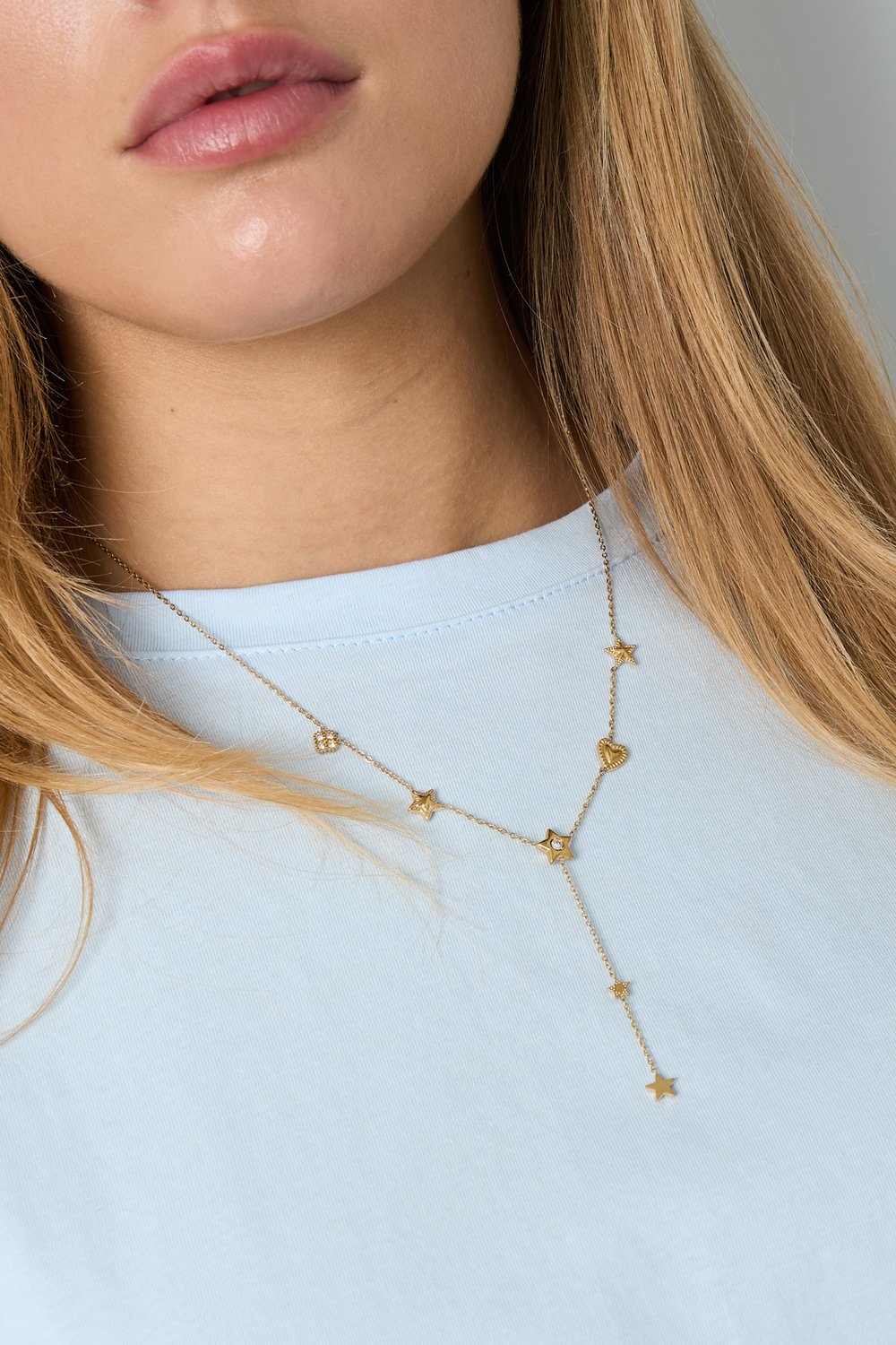 Gold lariat necklace featuring small star and heart charms worn by a woman against a light blue top.