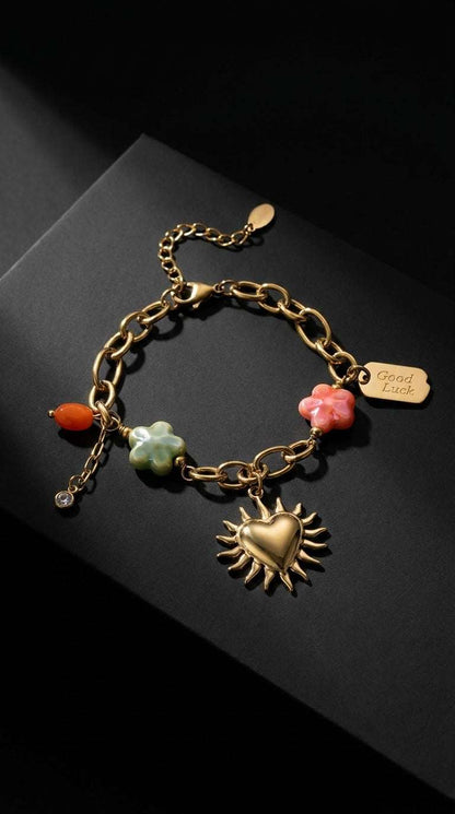 Gold charm bracelet featuring a sun-heart pendant, colorful flower charms, and a "Good Luck" tag on a dark background.