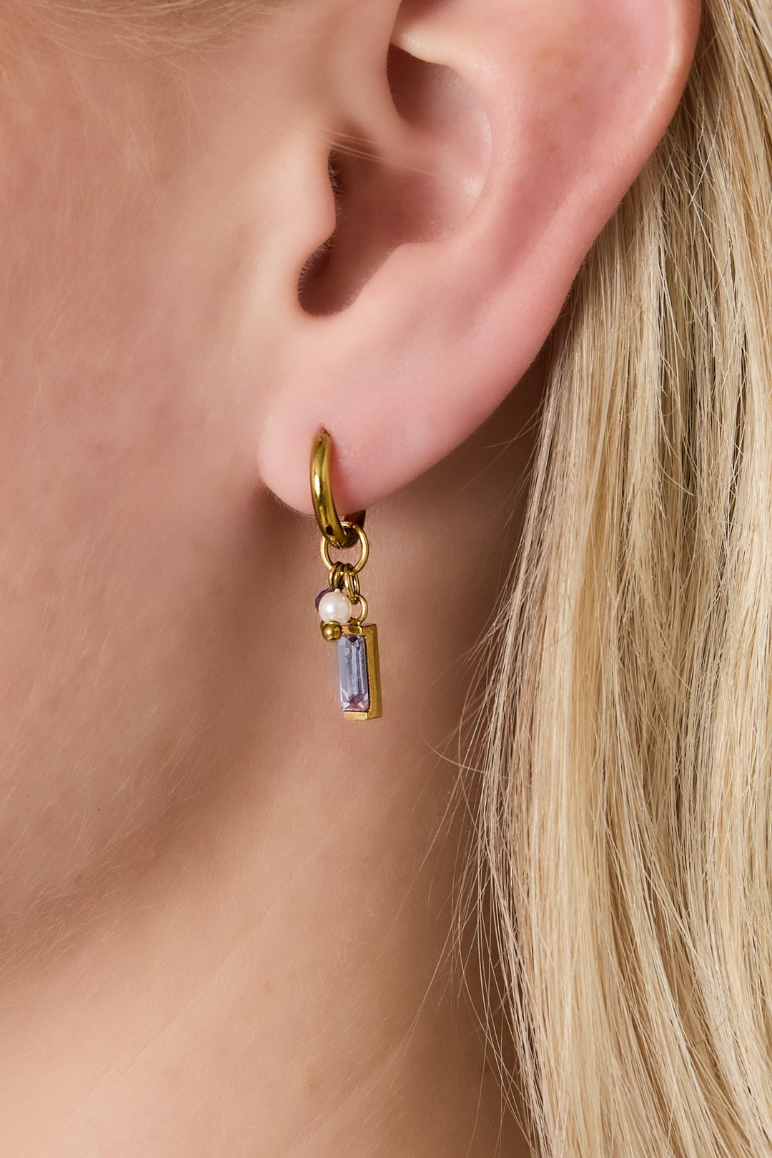Gold hoop earring with lilac rectangular gemstone, pearl, and purple bead worn on woman's ear with blonde hair