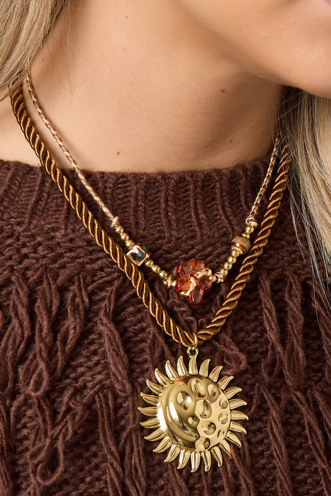 Close detail of layered necklaces on model, highlighting the textured gold sun pendant on twisted brown rope alongside a beaded floral necklace.