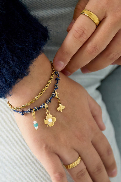 Close-up of hand wearing blue and gold cotton chain bracelet adorned with dangling gold charms including flower, bee, and beads, styled with gold rings and navy blue sweater.