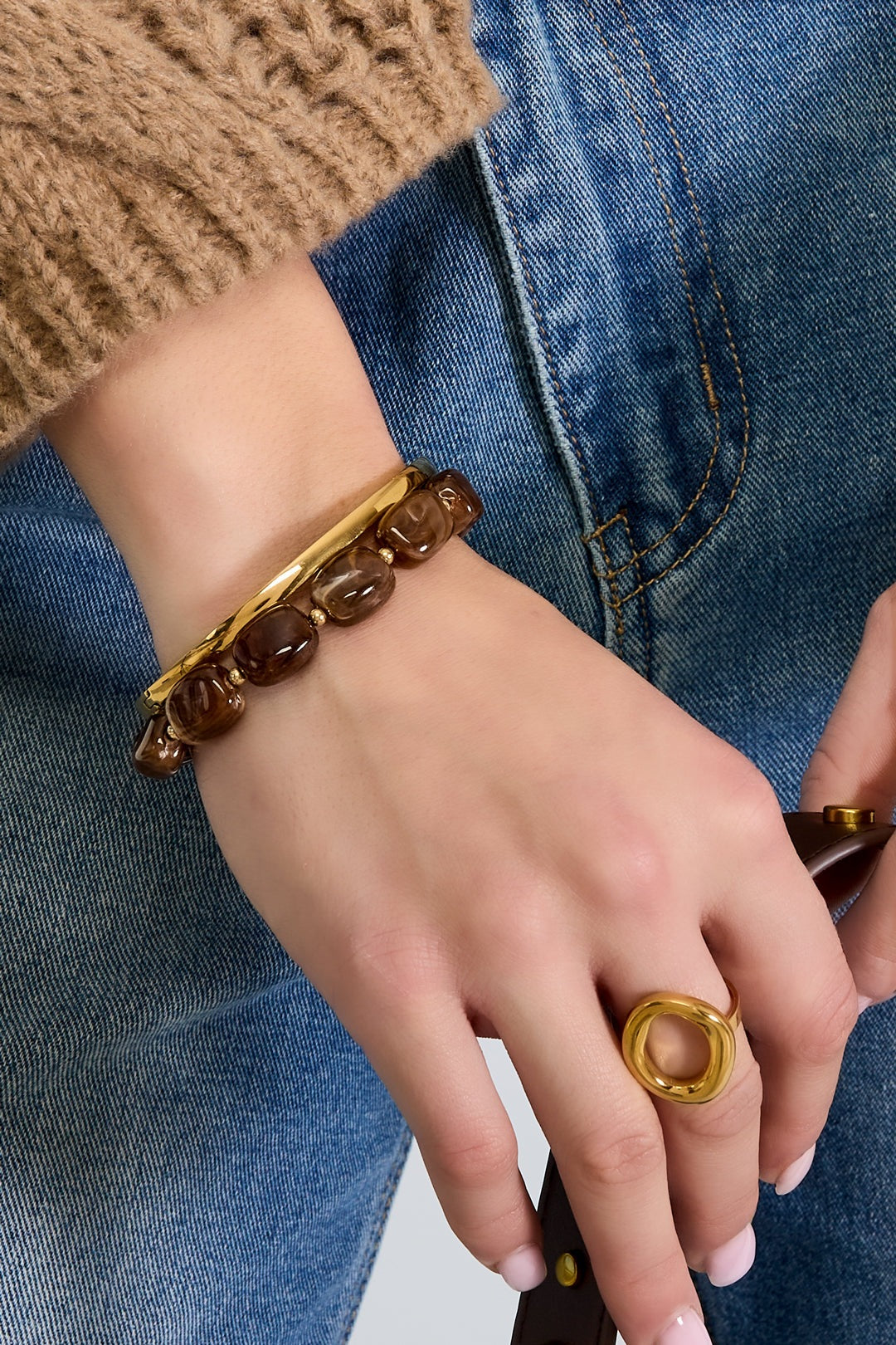 Model's wrist adorned with chocolate brown acrylic beaded bracelet paired with gold ring and bangle, wearing blue jeans and beige sweater