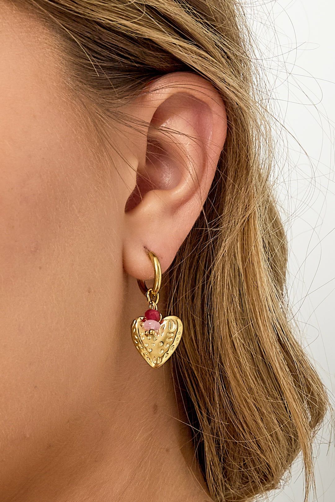 Side view close-up of woman’s ear showcasing single textured gold heart drop earring with pink and red beads
