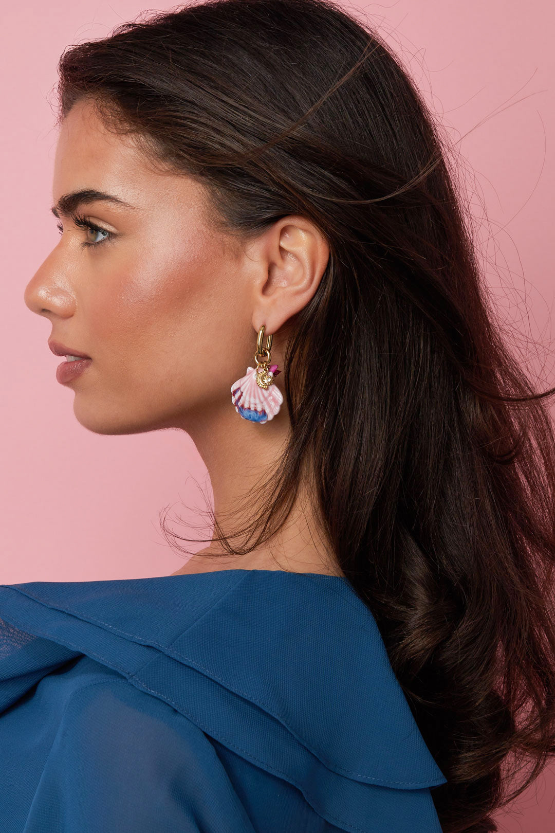 Side profile of woman wearing gold hoop earrings featuring hand-painted pink and blue seashell charms, with a pink background
