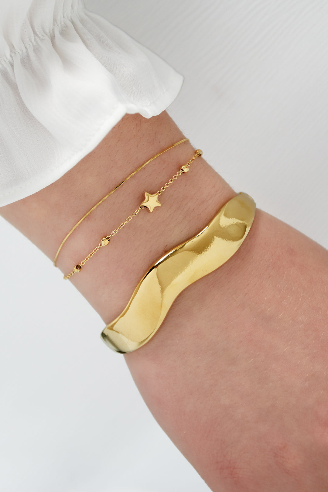 Close-up view of wrist wearing two gold bracelets; one delicate star charm bracelet paired with a smooth, wavy polished gold bangle and white sleeve.
