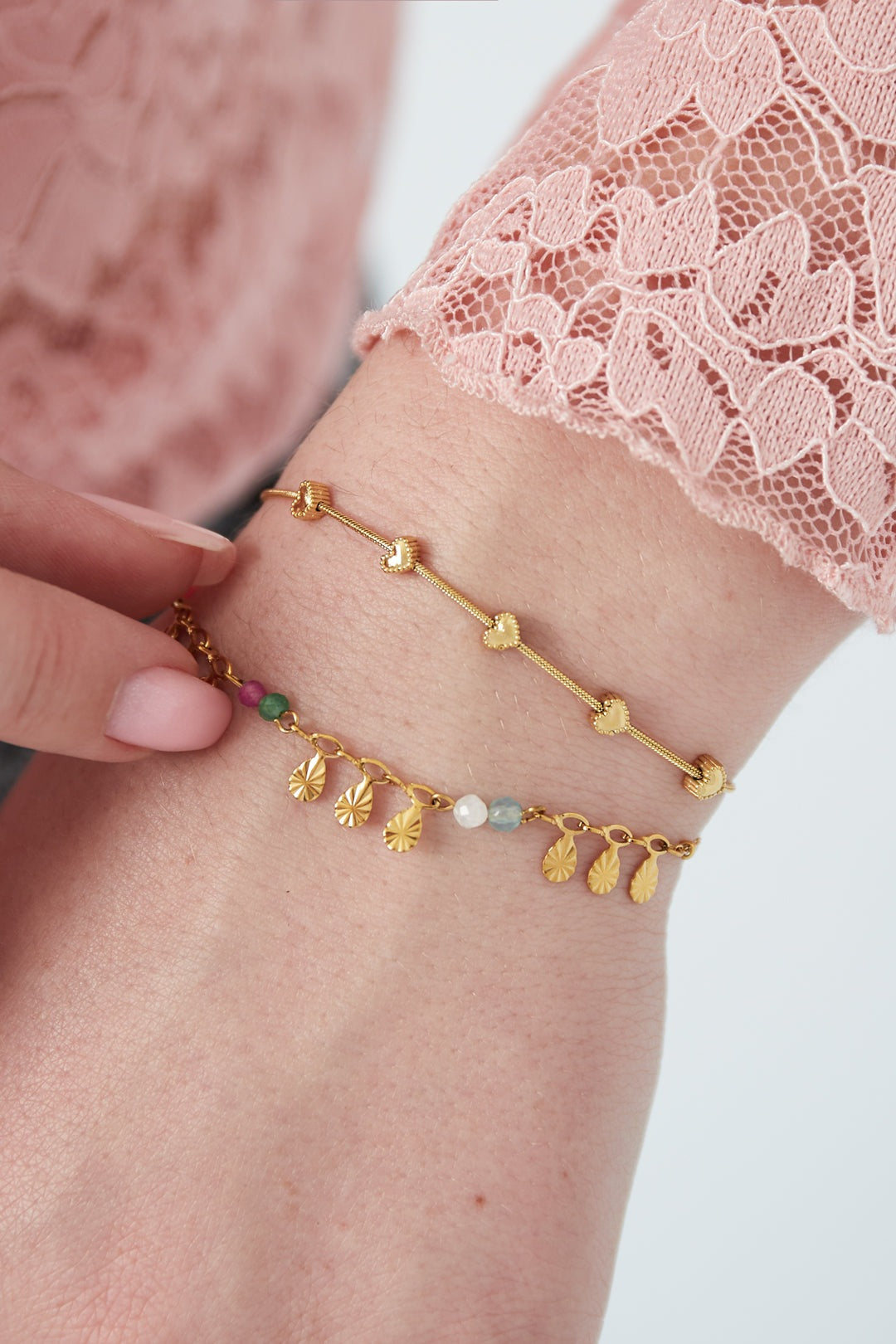 Close-up of a wrist wearing gold heart charm bracelet paired with a colorful charm bracelet, shown with light pink lace sleeve