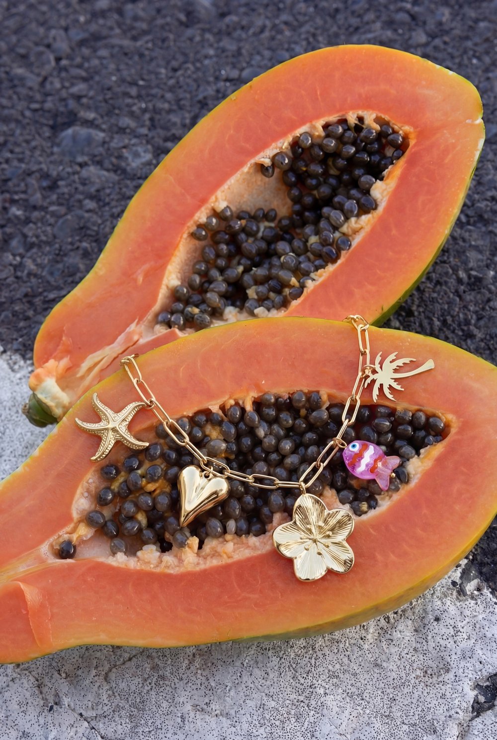 Gold link bracelet with starfish, heart, flower, palm tree, and fish charms resting on sliced papaya.
