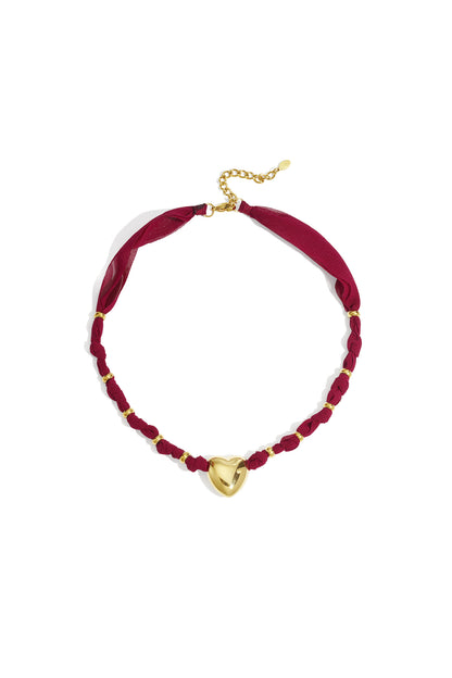 Gold heart pendant necklace with burgundy braided fabric and gold accents on white background
