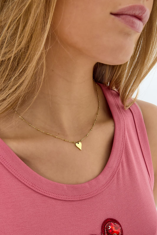 Close-up side view of the gold heart pendant necklace worn by woman in a pink top, showcasing the necklace’s delicate beaded chain.