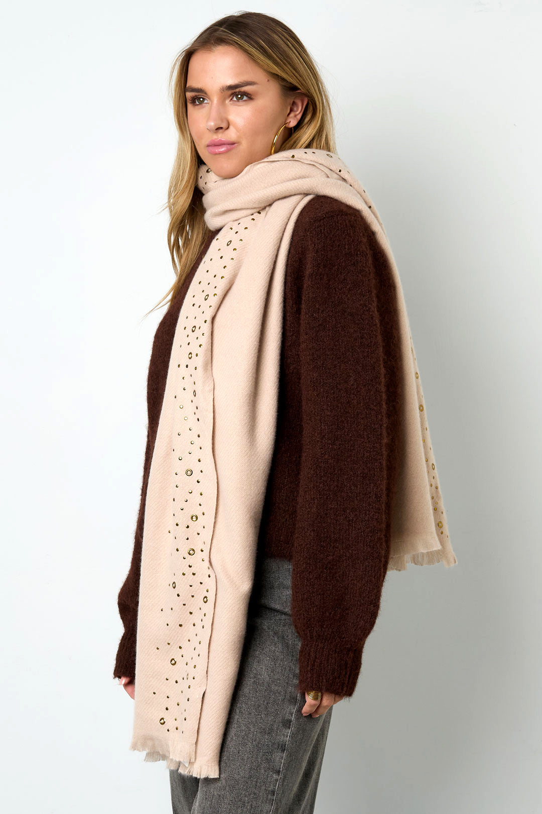 Side view of woman wearing cream studded scarf around neck over brown sweater and gray pants