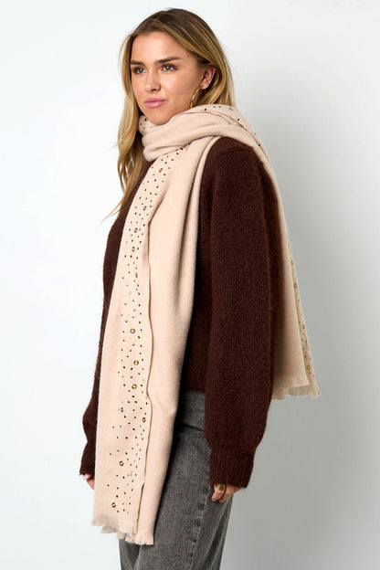 Side view of woman wearing cream studded scarf around neck over brown sweater and gray pants