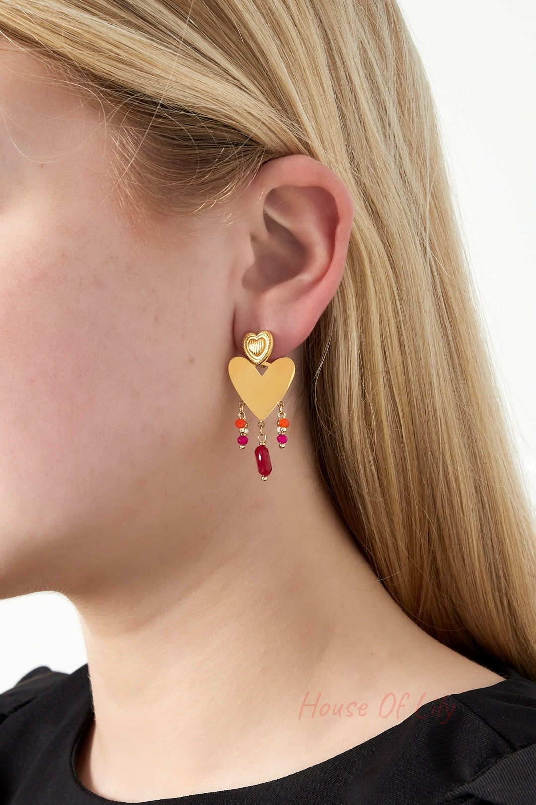 Model wearing gold-tone heart drop earring showing vibrant pink, orange, and red beads dangling below textured smaller heart stud with natural blonde hair and black top.