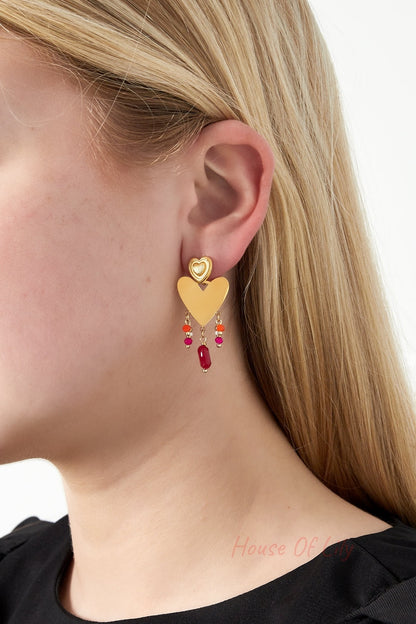 Model wearing gold-tone heart drop earring showing vibrant pink, orange, and red beads dangling below textured smaller heart stud with natural blonde hair and black top.