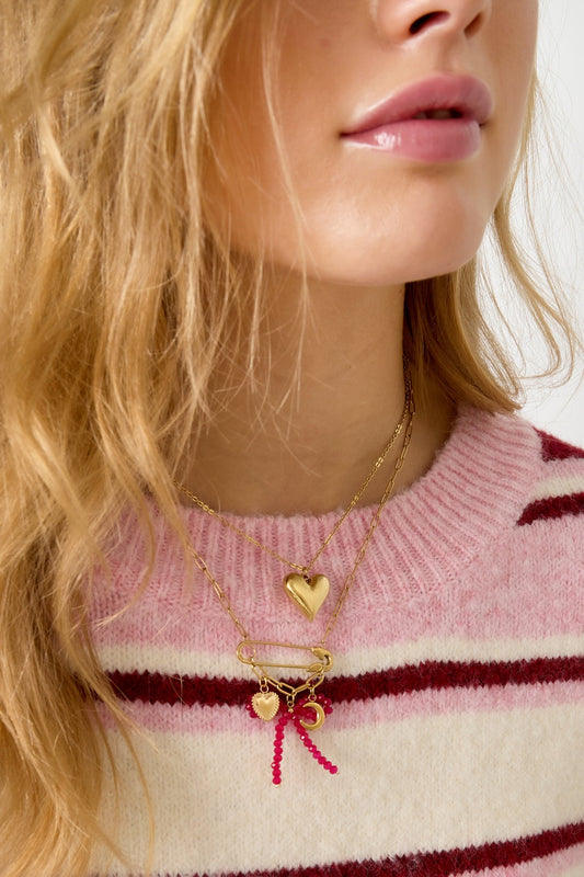 Close-up of woman wearing layered gold-tone necklaces including a safety pin pendant necklace with red beads and heart and moon charms