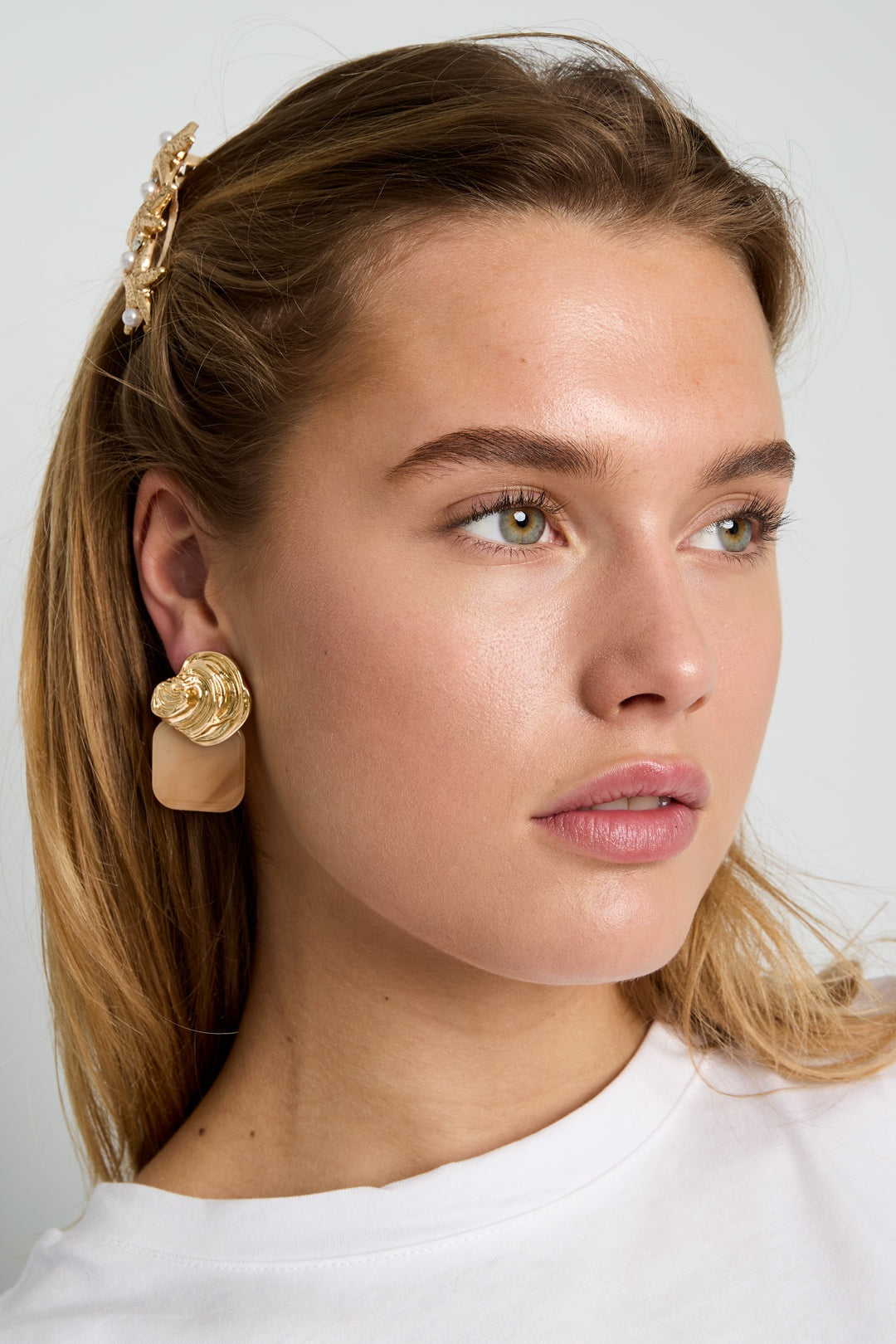 Model wearing Gilded Glitch Hoops: gold-toned, abstract textured studs paired with a square, smooth beige-toned pendant.