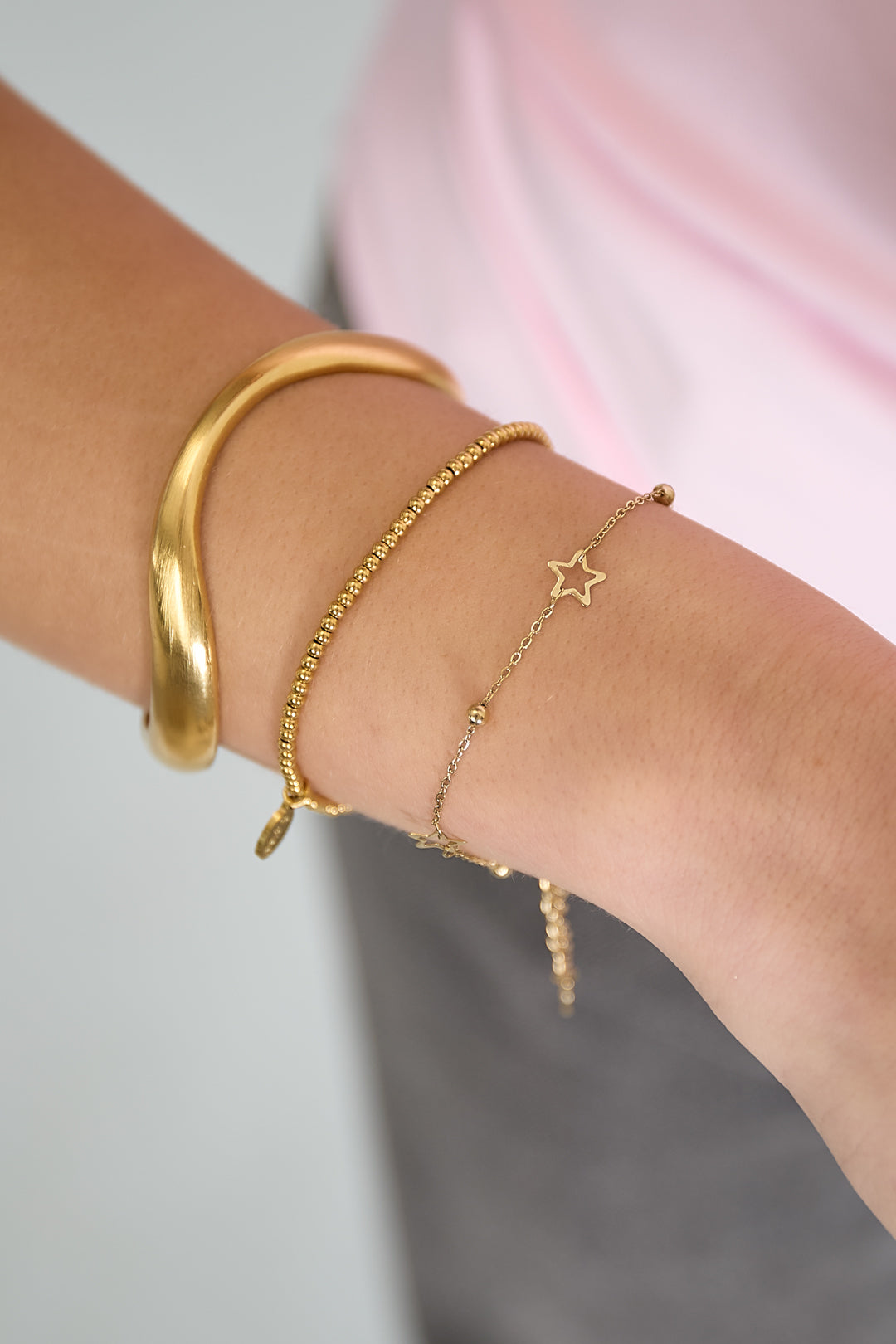 Close-up of a wrist wearing gold star chain bracelet stacked with other gold bracelets and pink top