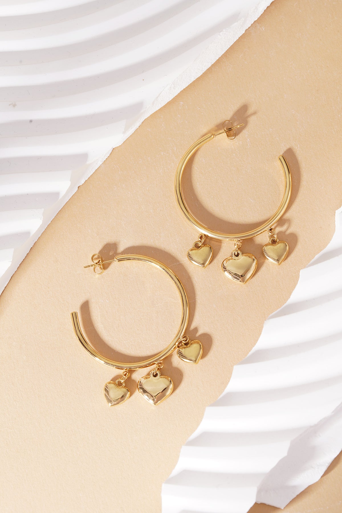 Gold hoop earrings with three dangling polished heart charms, displayed on a textured beige and white background.
