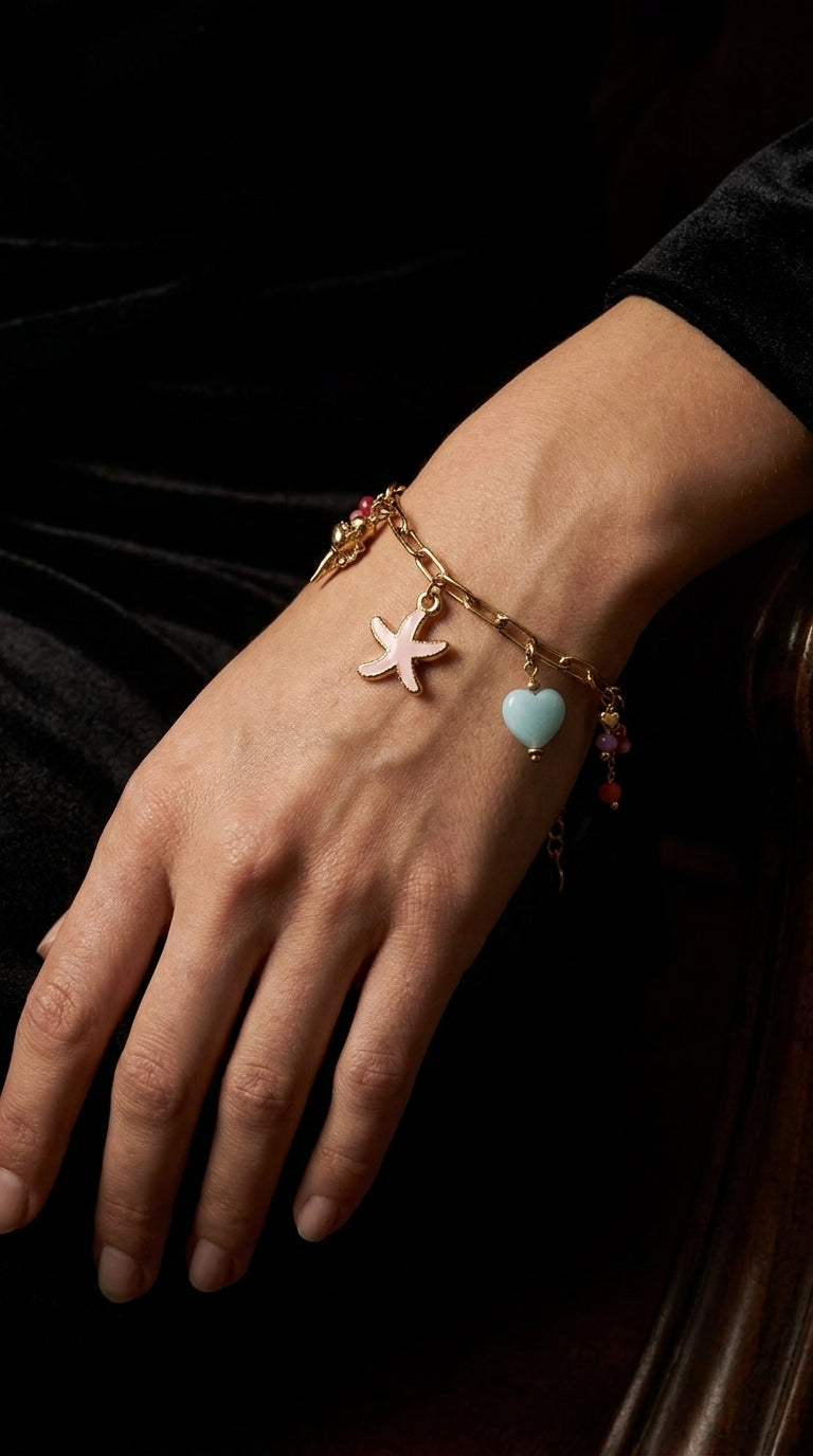 Coastal Charm gold chain bracelet with pink starfish and light blue heart charms, worn on a hand against black velvet.