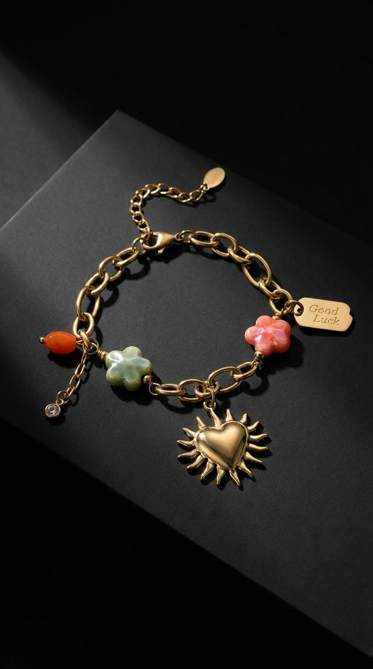 Gold charm bracelet featuring a sun-heart pendant, colorful flower charms, and a "Good Luck" tag on a dark background.