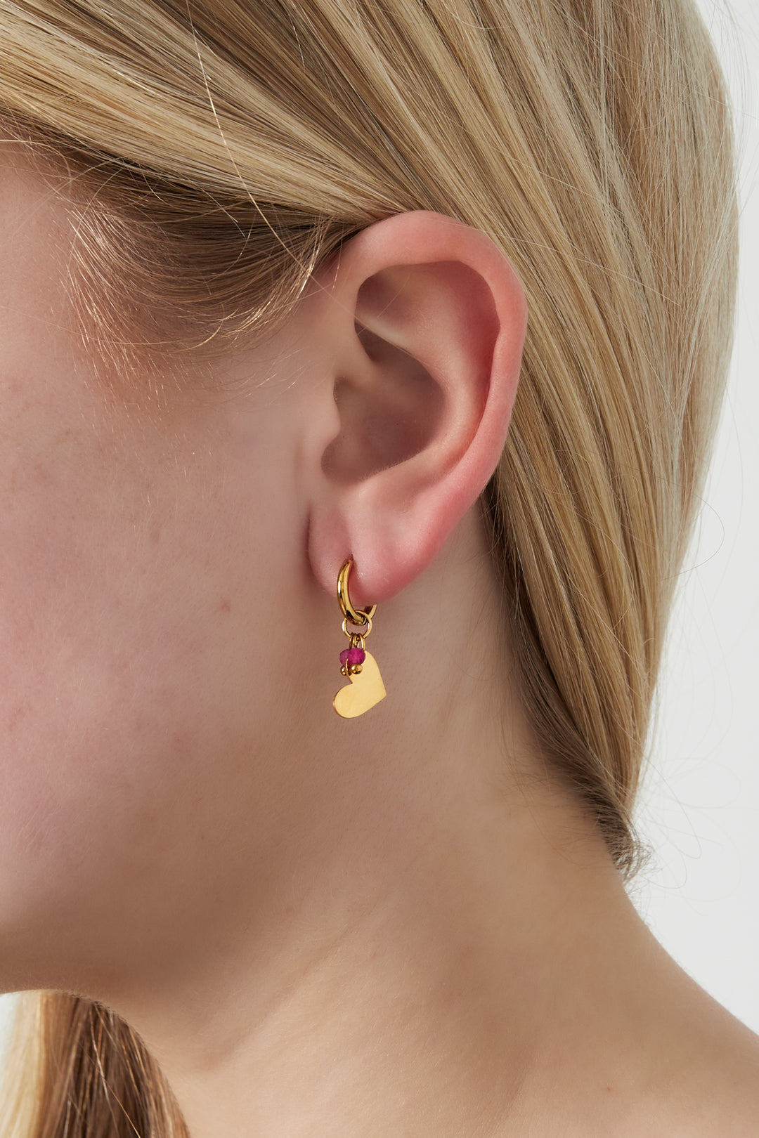 Close-up view of a woman wearing one gold hoop earring with a heart charm and small pink beads against a light background.