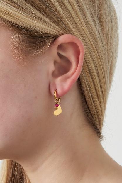 Close-up view of a woman wearing one gold hoop earring with a heart charm and small pink beads against a light background.