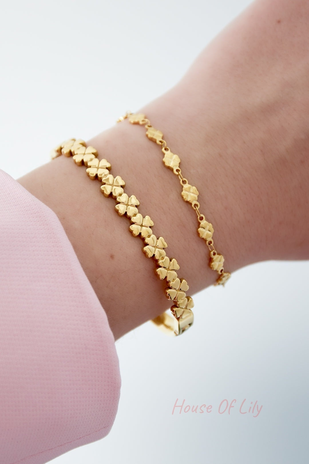 Close-up of wrist wearing two gold clover bracelets, one bangle and one chain style, paired with a pink sleeve