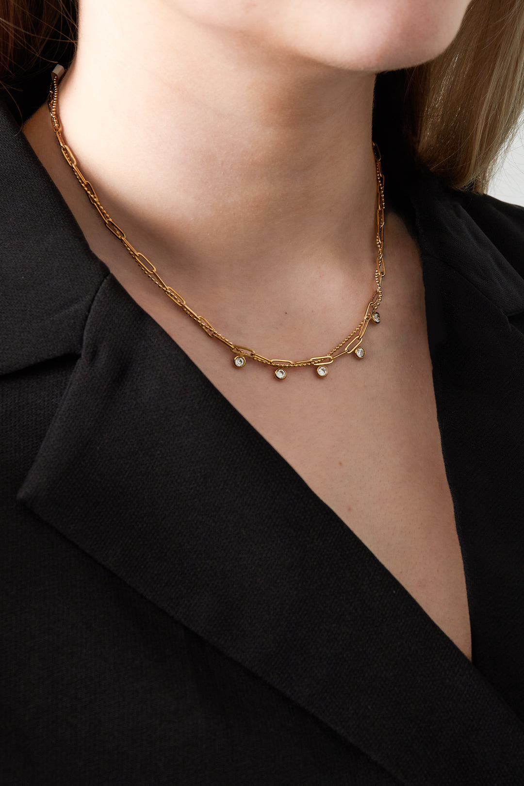 Model wearing the layered necklace with gold rectangular links and sparkling gemstone charms paired with a black blazer.