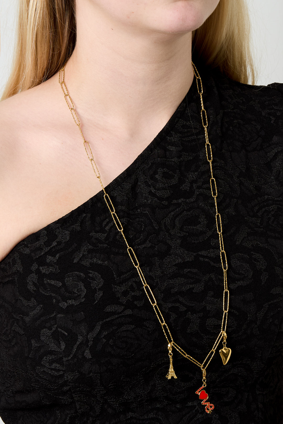 Gold elongated link chain necklace worn on a woman with black textured top, styled with small dangling charms