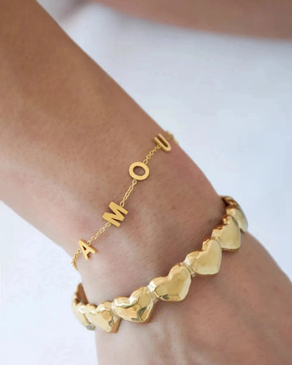 Two gold bracelets on a wrist: a delicate chain spelling "AMOUR" and a chunky bangle of polished, linked hearts.