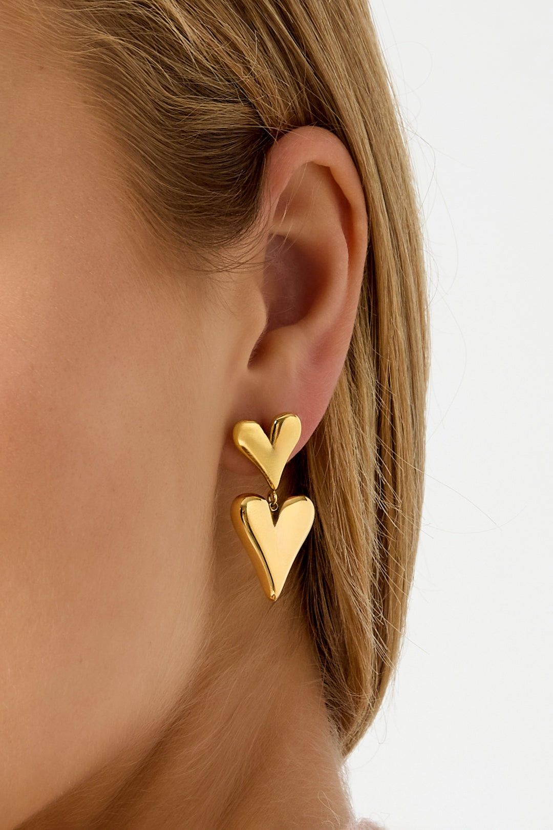 Side view close-up of woman wearing gold double heart drop earrings against white background
