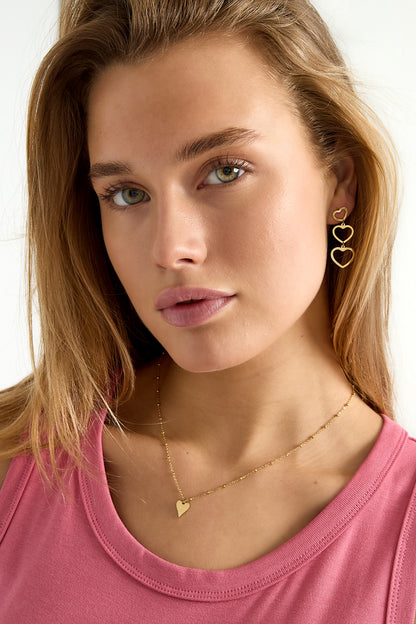 Woman wearing a youthful gold heart pendant necklace paired with matching hanging heart-shaped earrings, styled with a pink top.