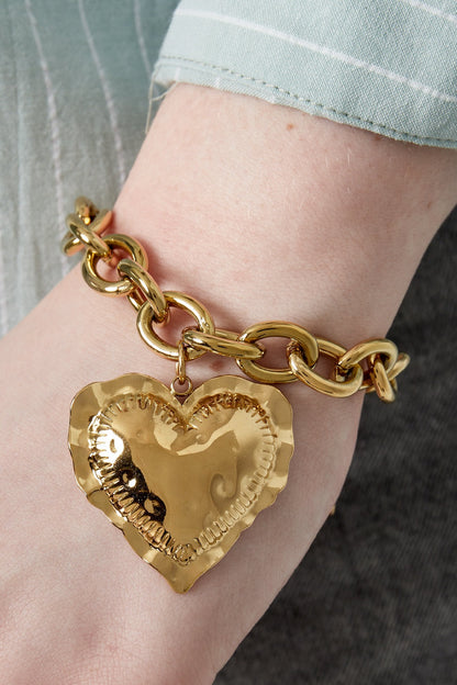 Close-up of gold chunky heart charm bracelet worn on wrist with soft green and gray outfit