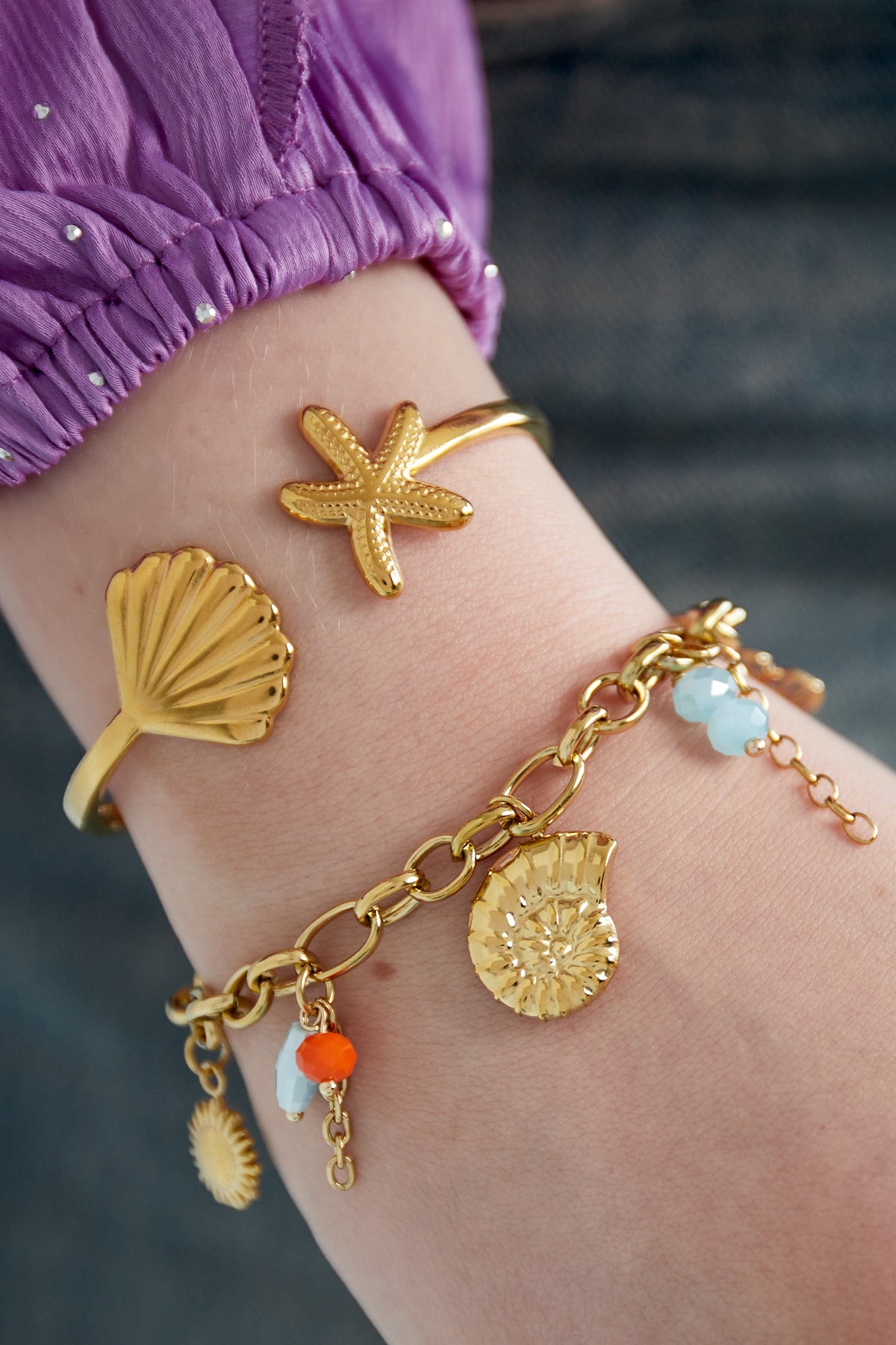 Close-up of bracelet worn with two gold sea motif bangles on wrist against purple sleeve and blue fabric