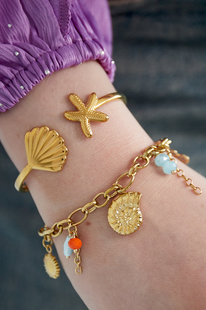 Close-up of bracelet worn with two gold sea motif bangles on wrist against purple sleeve and blue fabric