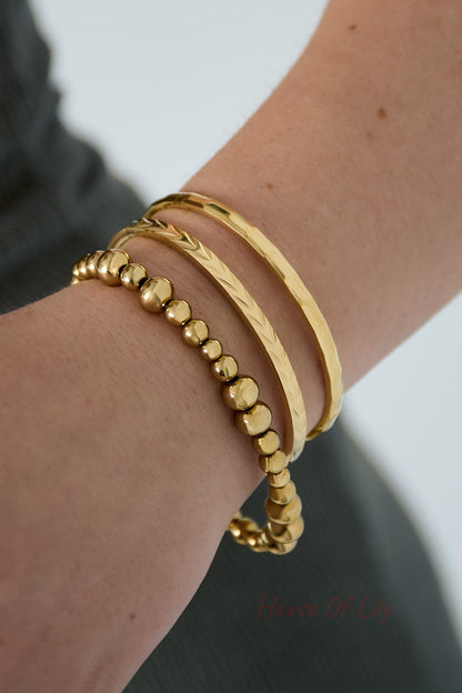 Close-up of a wrist wearing three gold bangles including geometric faceted bangle, textured band, and beaded bracelet