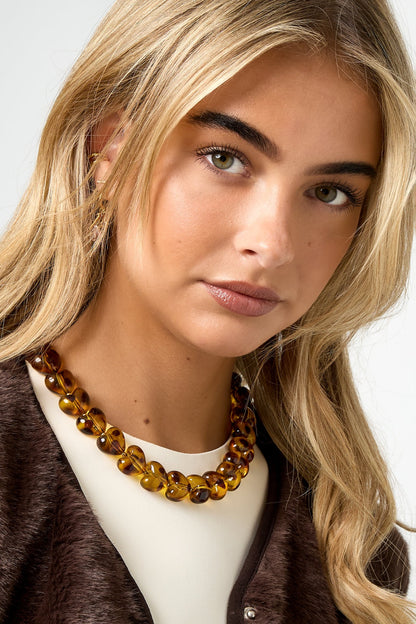 Close-up front view of woman wearing amber heart bead necklace paired with cream top and brown vest