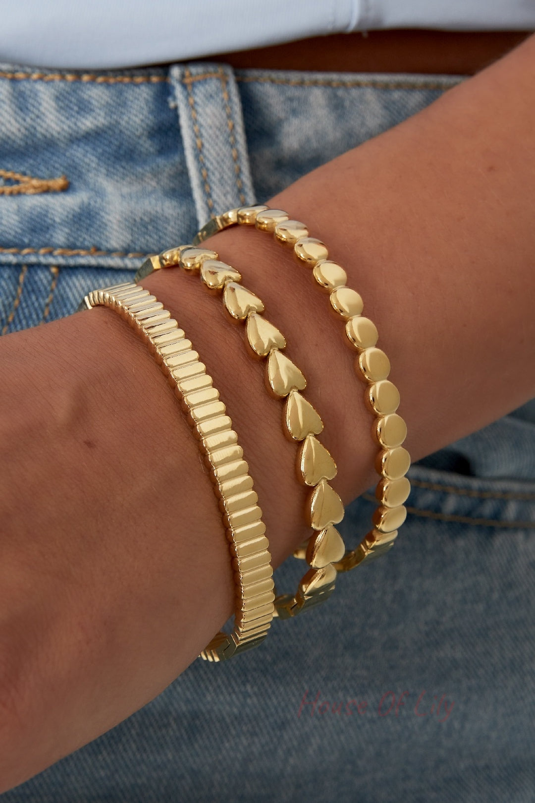 Close-up of three gold bracelets stacked on woman's wrist, including the textured bangle, heart and round shape bangles