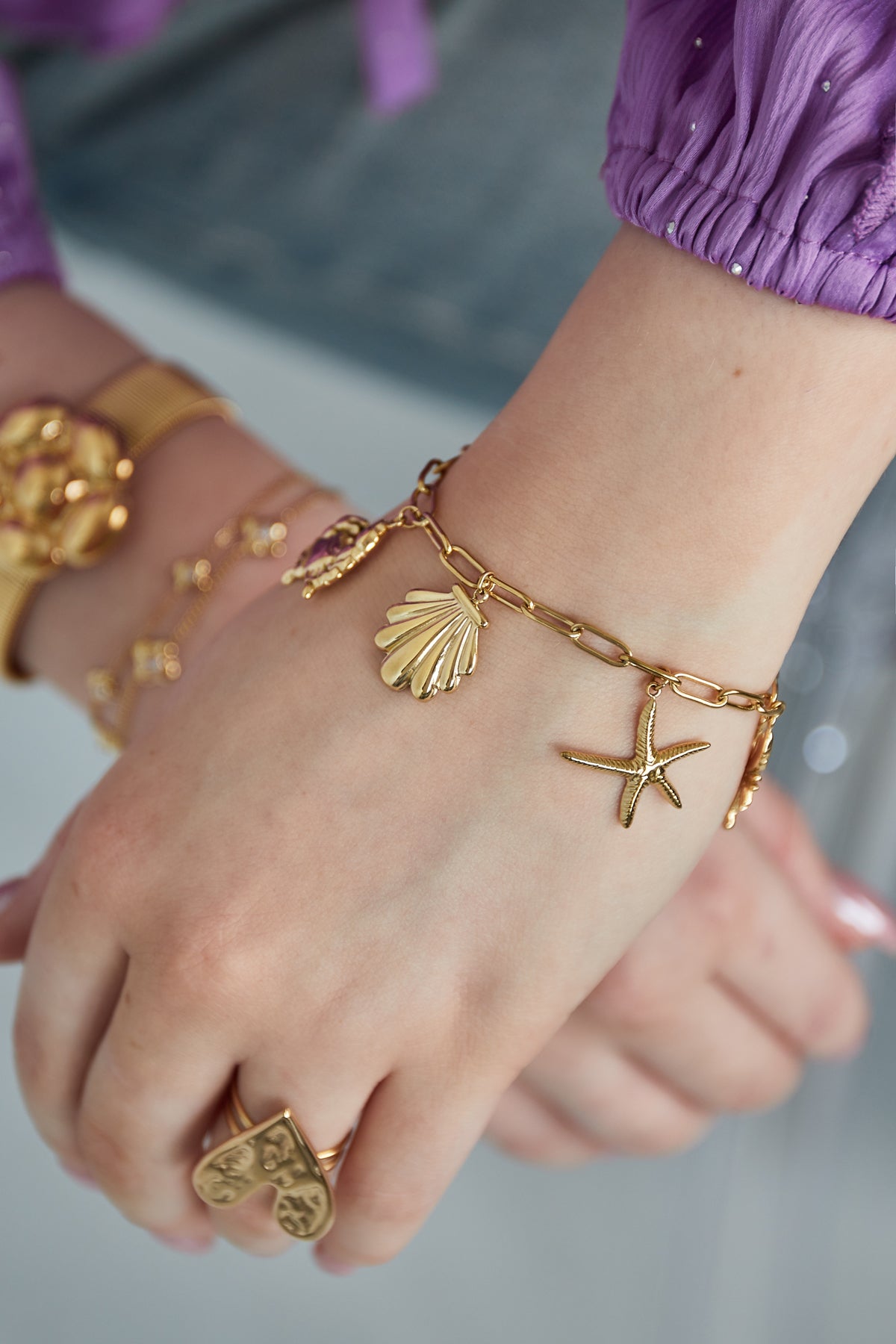 Close-up of gold-tone seaside charm bracelet worn on wrist with purple top and heart ring