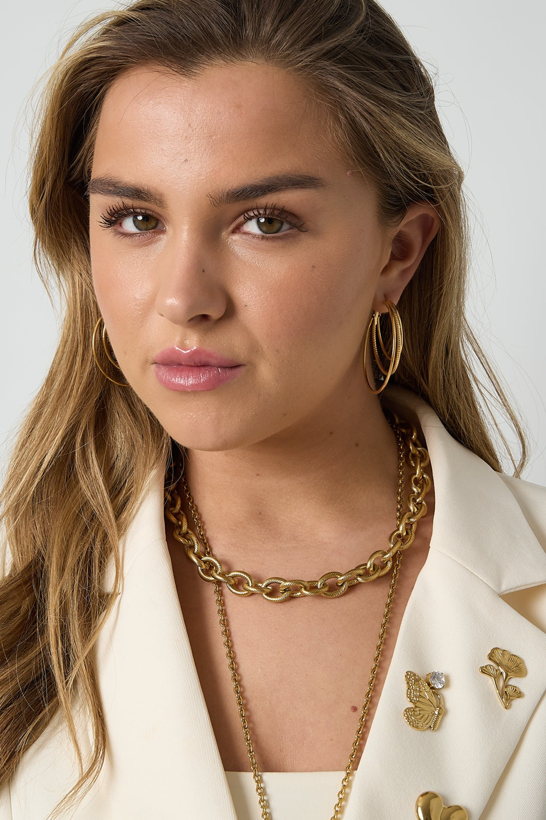 Woman wearing gold triple layered hoop earrings paired with gold chain necklaces and cream blazer adorned with floral and butterfly pins