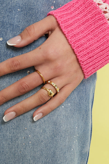 Woman's hand wearing three gold rings including a minimalist gold heart ring, styled with pink knitted sleeve and denim jeans
